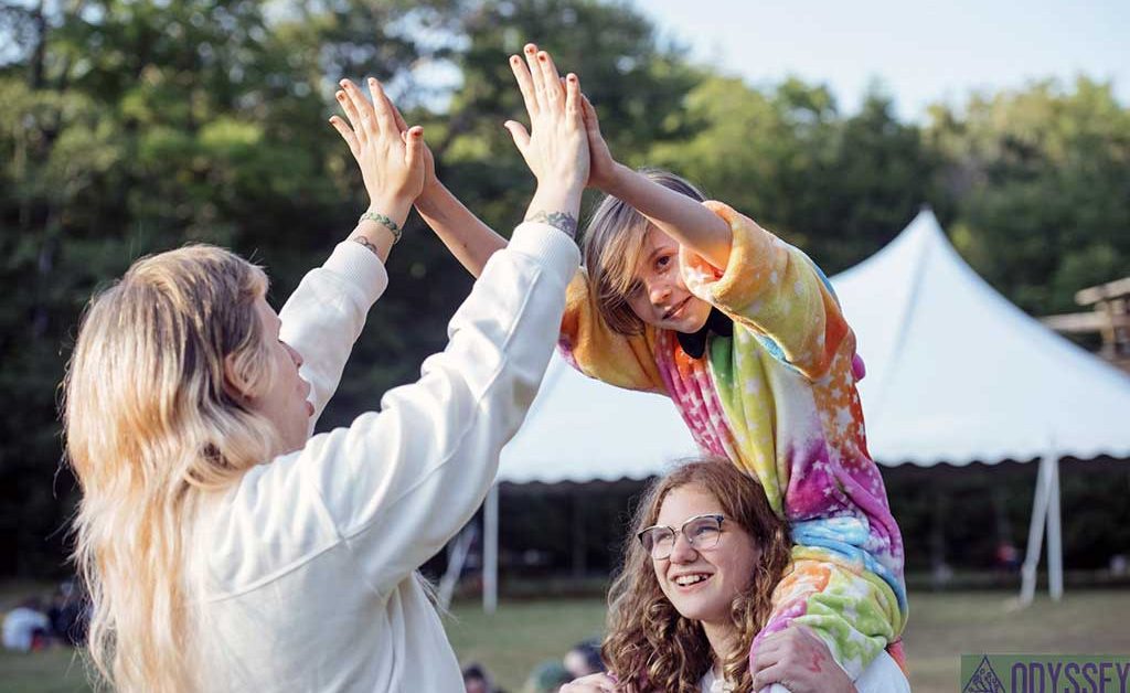 Young camper gives counselor a high five while sitting on shoulders of another camper.