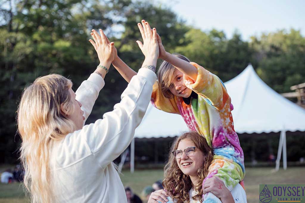 Young camper gives counselor a high five while sitting on shoulders of another camper.