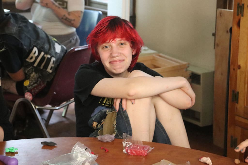 Smiling teen girl with dyed red hair, sitting with hands wrapped around knees in art hut