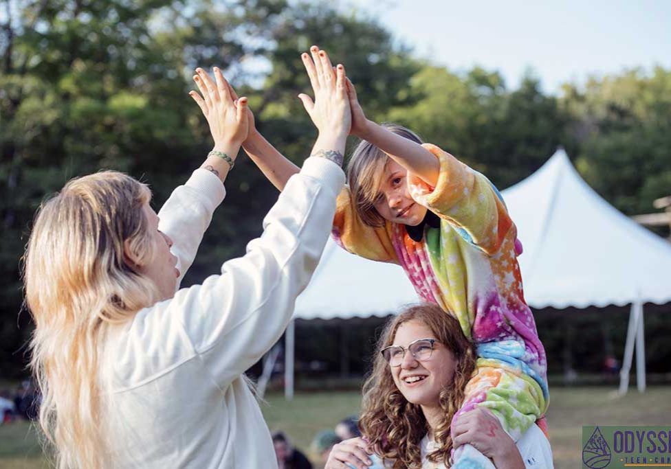 Young camper gives counselor a high five while sitting on shoulders of another camper.
